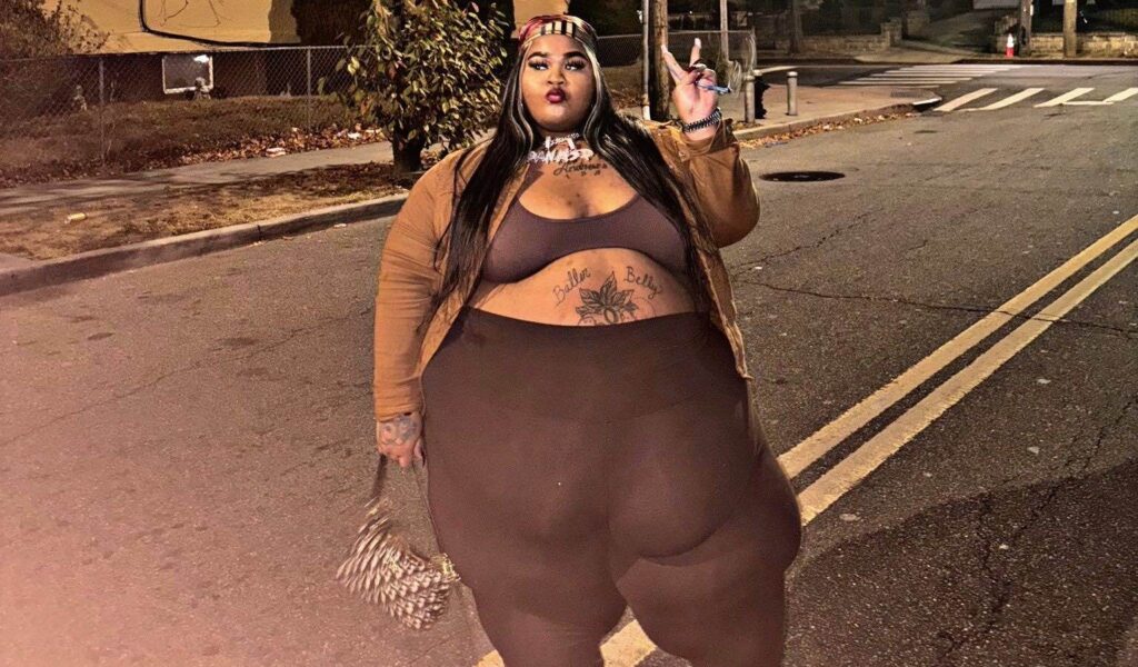 Rapper Dank DeMoss posing confidently on a street, wearing a brown jacket and leggings, with visible tattoos and a stylish handbag, reflecting themes of body positivity amid discussions of weight discrimination in a lawsuit against Lyft.