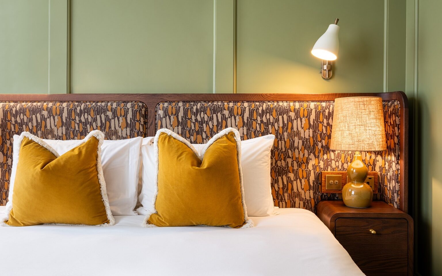 olive green panelled wall with patterned fabric headboard and mustard yellow cushions in Mollies Manchester