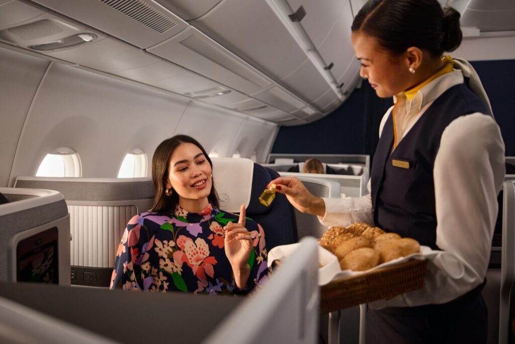 a woman sitting in an airplane with a tray of bread and a woman holding a bottle of oil