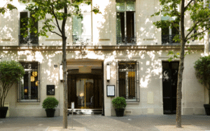 Pale facade of Le Narcisse Blanc Hotel, dappled with sunlight from trees lining the boulevard